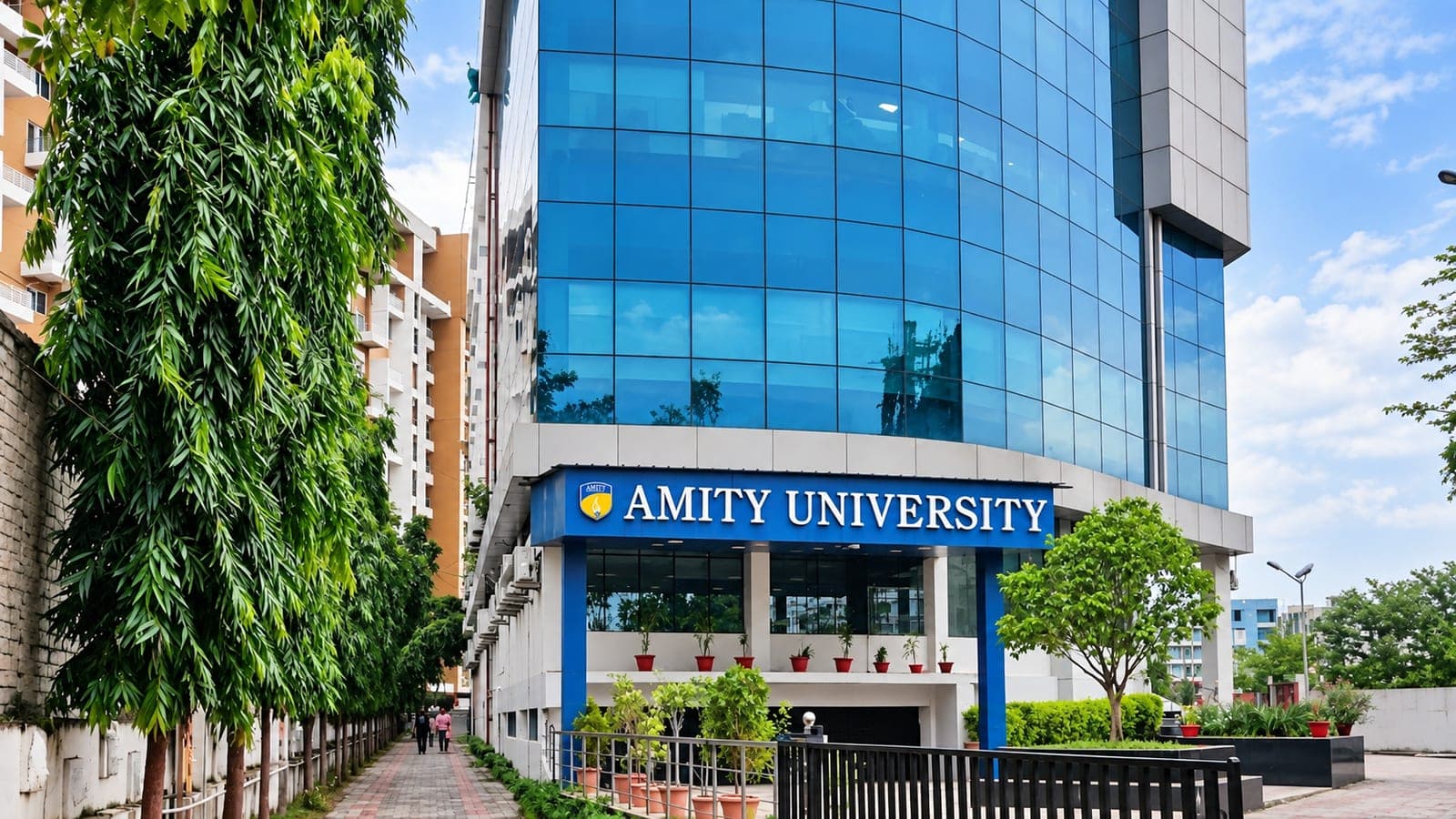 The Amity University Patna campus: a blue glass academic tower with the university's name above the entrance, at the end of a tree-lined walk.